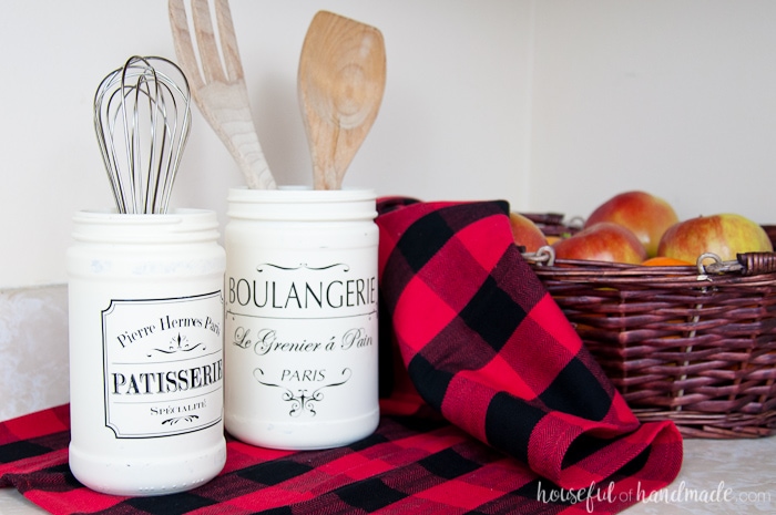 set of two white painted bakery jars shown on buffalo plaid next to basket.