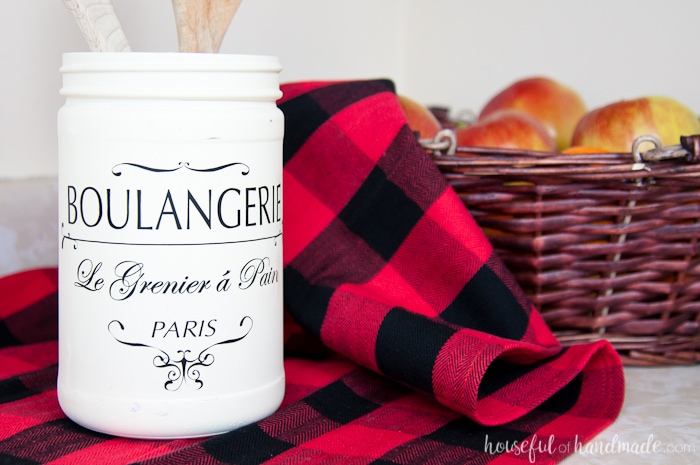 white painted jar with french quote shown on red buffalo plaid next to basket.