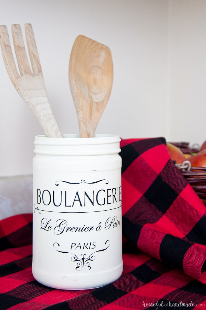 Refinished kitchen jar painted white shown with two kitchen utensils on buffalo plaid cloth.